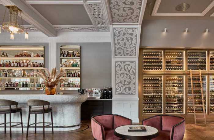 Luxus Hotelbar: Links die elegante Bar aus wertvollem Stein, rechts ein Einblick in die Auswahl an Wein & Whisky, darüber die ikonische Stuckdecke.