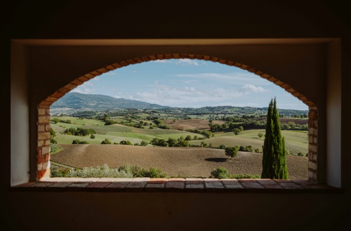 Panoramablick auf eine hügelige Landschaft mit grünen und braunen Feldern, einzelnen Bäumen und fernen Hügeln, gesehen durch ein gemauertes Bogenfenster.