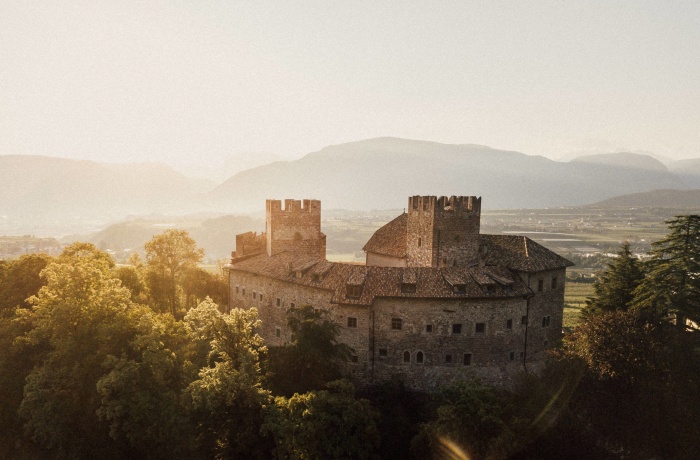 Mittelalterliches Schloss aus Stein mit Türmen und Zinnen, umgeben von Bäumen, im warmen Licht der untergehenden Sonne mit Blick auf das Tal.