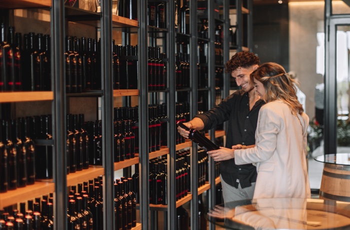 Ein Paar steht in einem modernen Weinkeller vor Regalen voller Weinflaschen und betrachtet gemeinsam eine Flasche.