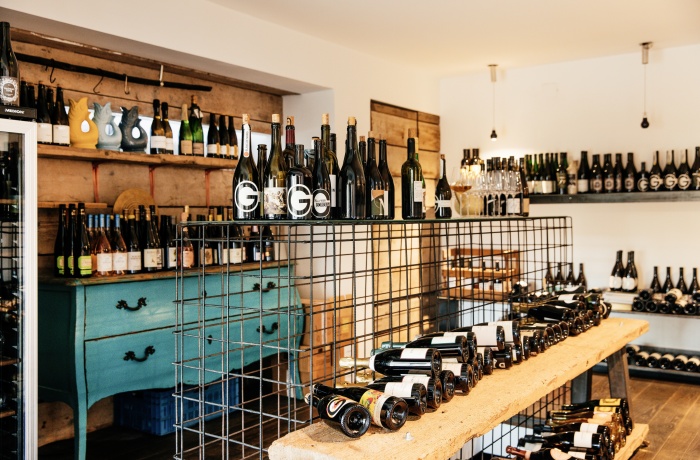 Innenansicht eines Weinladens mit Regalen und Tischen voller Weinflaschen, darunter eine alte türkisfarbene Kommode als Blickfang.