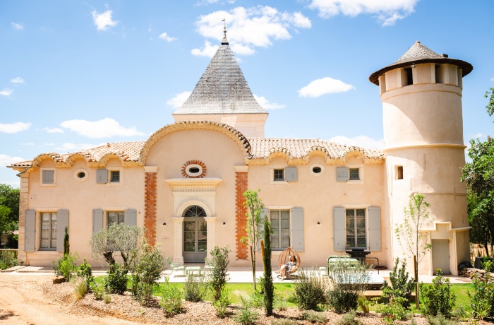 Helles südfranzösisches Schloss mit rundem Turm, grauen Fensterläden und gepflegtem Garten unter blauem Himmel.
