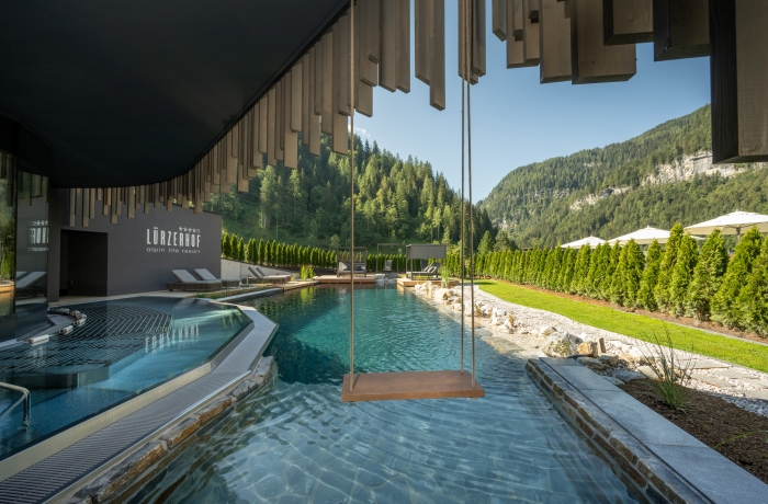 Außenpool eines Wellnesshotels mit Schaukel über dem Wasser und Bergpanorama im Hintergrund.

