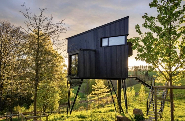 Futuristisches Glamping-Baumhaus auf Stelzeln, umgeben von leichter Bewaldung und Wiesen