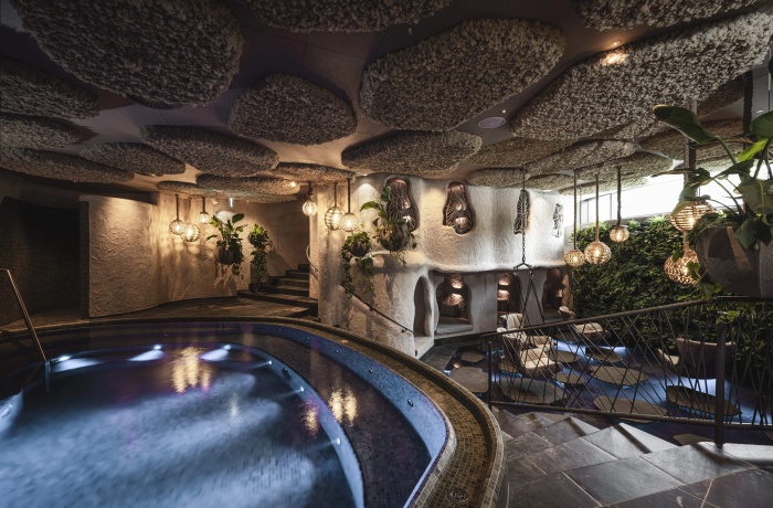 Stimmungsvoll beleuchteter Spa-Innenbereich mit einem runden Whirlpool, organisch geformten Wänden und gemütlichen Nischen zum Entspannen. Die Decke ist mit wolkenartigen Strukturen gestaltet, es hängen dekorative Lampen aus Naturmaterialien und rundherum sind viele Pflanzen integriert.