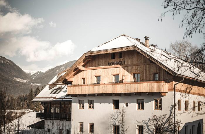 Ein modernisiertes, alpin-traditionelles Gasthaus, im Hintergrund verschneite Berge und Tannenwälder
