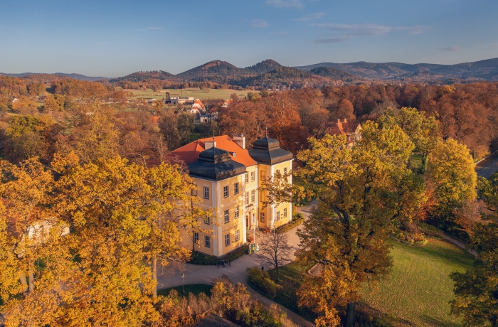 Luftaufnahme eines gelben Barockschlosses mit rotem Dach, umgeben von herbstlich gefärbten Bäumen und Parkanlagen, im Hintergrund eine hügelige Landschaft mit Bergen unter blauem Himmel.