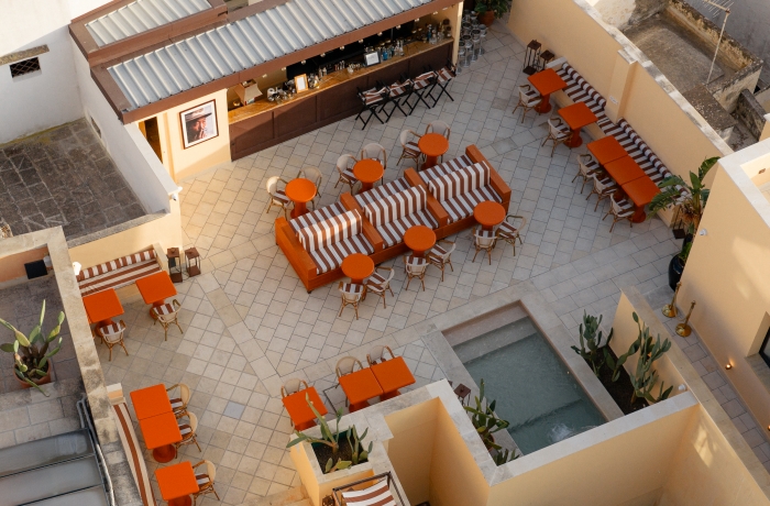 Der stilvolle Innenhof des Palazzo Presta lädt mit seiner modernen Bar, orangefarbenen Sitzmöbeln und mediterranem Ambiente zum Verweilen ein. Klare Linien, warme Farbtöne und ein kleiner Wasserbereich schaffen eine entspannte Atmosphäre im Herzen Apuliens.