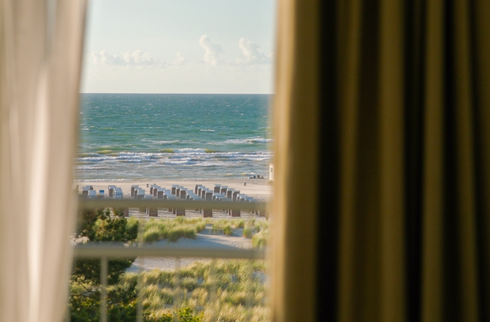 Blick aus einem Hotelzimmer auf die Ostsee mit Sandstrand, Strandkörben und sanften Dünen vor dem weiten Horizont.