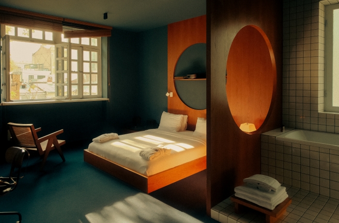 Modernes Hotelzimmer mit niedrigem Doppelbett, warmem Sonnenlicht durch große Fenster, farblich abgesetzter Wandgestaltung und offen integriertem Badbereich mit gefliester Badewanne.