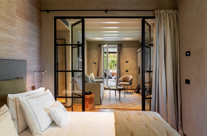 Modernes Hotelzimmer mit großem Bett im Vordergrund, Glas-Schiebetüren zum gemütlichen Wohnbereich mit Sofa und Sessel sowie Zugang zu einer Terrasse mit Liegestuhl.