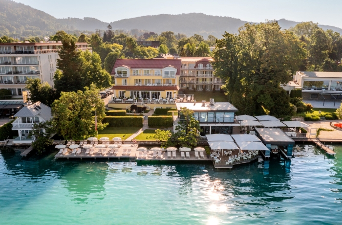 Luftaufnahme des Seehotel Dr. Jilly direkt am türkisblauen Seeufer mit Sonnenterrasse, Liegen und bewaldeten Bergen im Hintergrund.