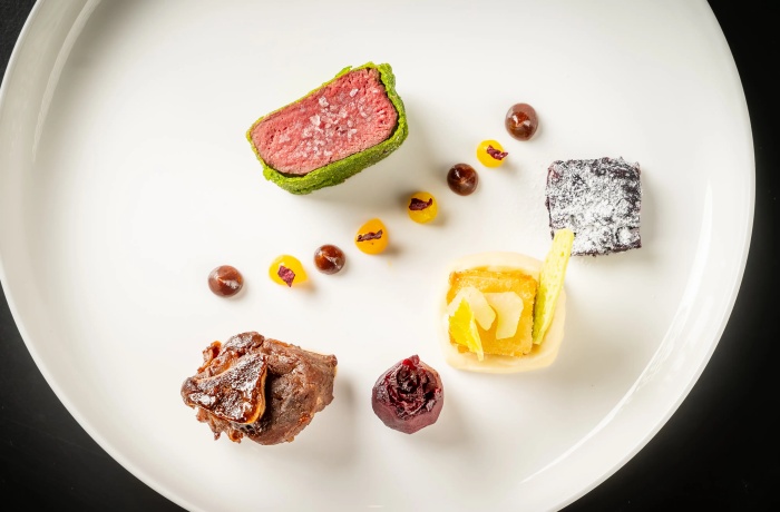 Kunstvoll angerichtetes Gourmetgericht auf weißem Teller: rosa gegartes Fleischstück in Kräuterkruste, begleitet von kreativen Gemüse-, Frucht- und Saucenelementen in moderner Anordnung.