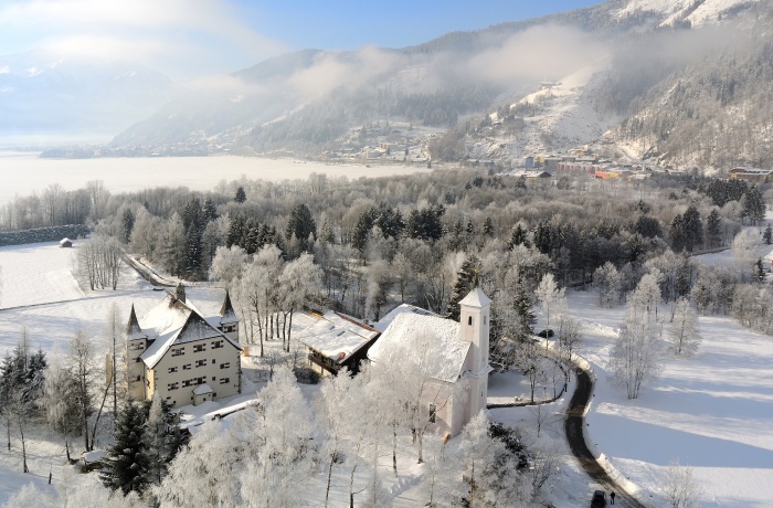 Winterliche Luftaufnahme von Schloss Prielau und einer Kirche, umgeben von verschneiten Bäumen und Bergen im Hintergrund.