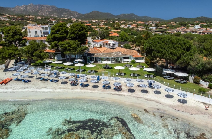 Luftaufnahme des The Pelican Beach Resort & SPA mit orangefarbenen Dächern, umgeben von Bäumen und Bergen; davor ein heller Sandstrand mit Liegen und Sonnenschirmen sowie türkisfarbenes, klares Wasser.