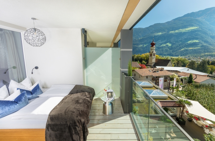 Modern eingerichtetes Hotelzimmer mit großem Doppelbett, Balkon und weitem Blick auf eine sonnige Berglandschaft und ein Dorf im Tal.