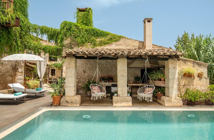 Moderne, mediterrane Villa mit Natursteinfassade, großer Terrasse und privatem Pool im Vordergrund. Sonnenliegen und eine überdachte Sitzlounge bieten entspannte Rückzugsmöglichkeiten. Der Himmel ist strahlend blau mit vereinzelten weißen Wolken, umgeben von schlichter Gartenbepflanzung und hohen Mauern für Privatsphäre.