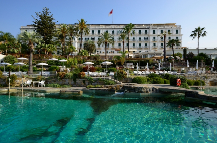 Elegantes Grandhotel mit mediterraner Gartenanlage, Palmen und luxuriösem Poolbereich direkt am klaren Wasser der Küste.