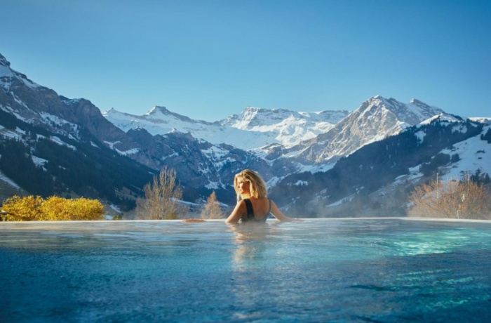 Frau entspannt im Infinity-Pool des The Cambrian Hotels, mit spektakulärem Blick auf schneebedeckte Berge und eine sonnige Winterlandschaft.