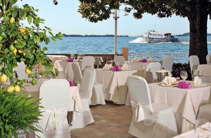 Hotelrestaurant mit weißen Tischen und Stühlen mit direkten Blick auf den Gardasee, ein Schiff fährt vorbei