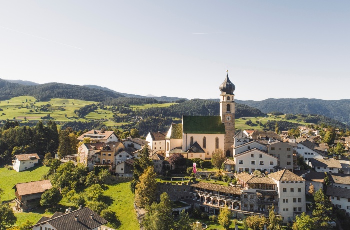 Luftaufnahme eines malerischen Dorfes in den Bergen mit dem Romantik Hotel Turm und der Kirche mit Zwiebelturm, umgeben von grüner Hügellandschaft.