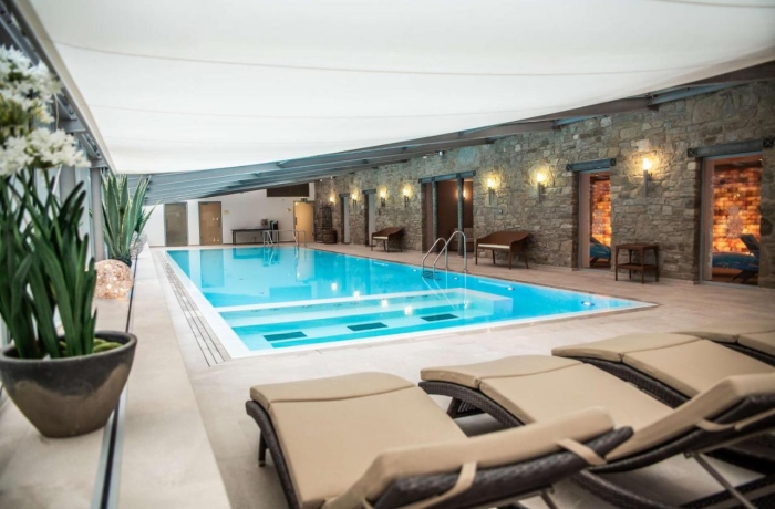 Eleganter Indoor-Poolbereich mit modernen Liegen, Natursteinwänden und entspannter Spa-Atmosphäre unter hellem Deckensegel.