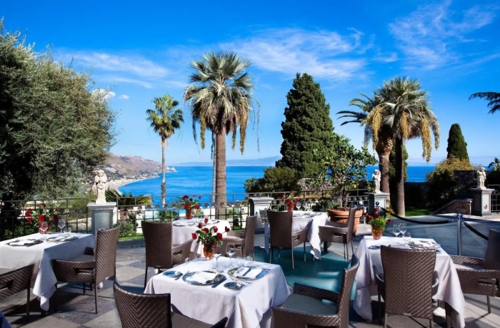 Hotels mit Gourmetrestaurant: Terrasse eines eleganten Hotelrestaurants mit weiß gedeckten Tischen, Meerblick, Palmen und mediterranem Flair unter strahlend blauem Himmel.