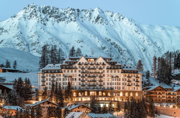 Das beleuchtete Carlton Hotel St. Moritz vor einer beeindruckenden Kulisse schneebedeckter Berge und winterlicher Landschaft in der Abenddämmerung.