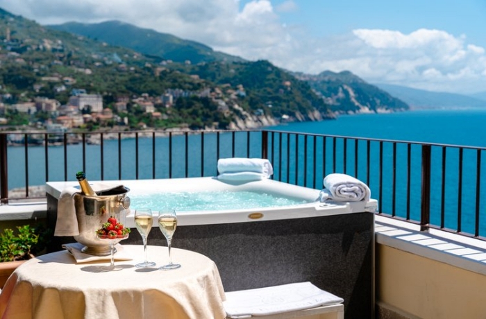 Der private Balkon im Hotel Excelsior Palace an der Portofino-Küste bietet einen malerischen Ausblick auf das Küstengebiet. Ein privater Whirlpool auf dem Balkon verspricht Entspannung mit Blick auf das atemberaubende Panorama. Im Vordergrund steht ein ansprechend gedeckter Tisch mit einem Sektkühler, zwei Sektgläsern und einer Etagere voller frischer Erdbeeren. Diese elegante und romantische Atmosphäre lädt zu unvergesslichen Momenten in einer der begehrtesten Regionen Italiens ein