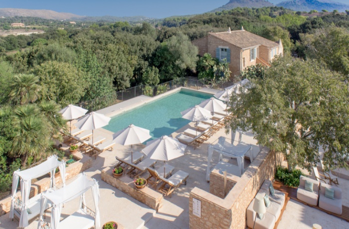 Historisches Hotel im traditionellen Architekturstil Mallorcas, mit viel freigelegter Steinfassade, einem Pool sowie Liegen und weißen Sonnenschirmen.
