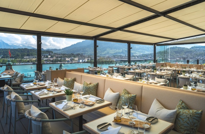 Hotels mit Gourmetrestaurant: Modern gedeckte Terrasse eines Hotelrestaurants mit Blick auf See und Berge, stilvollen Sitzpolstern und elegantem Ambiente unter Sonnenschutz.