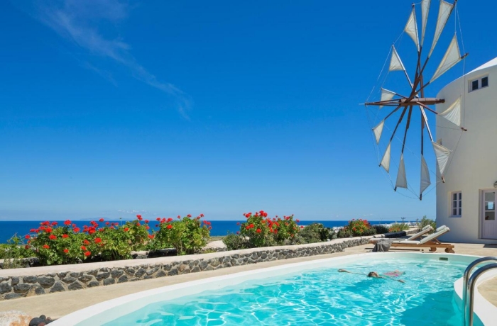 Außenpool mit türkisblauem Wasser in einer mediterranen Ferienanlage, umgeben von Blumen, mit Blick auf das Meer und eine Windmühle im Hintergrund.
