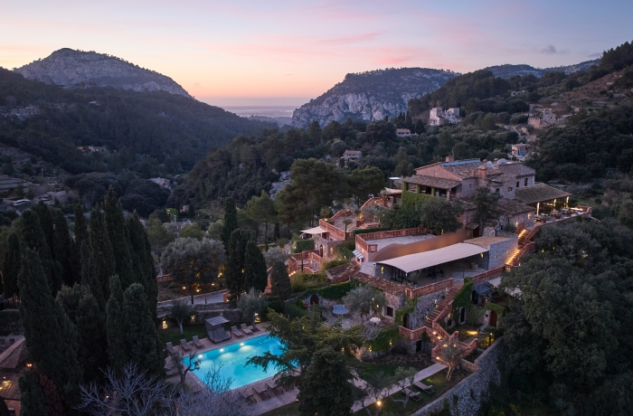 Malerisches Luxushotel in den Bergen Mallorcas bei Sonnenuntergang, eingebettet in eine üppige mediterrane Landschaft. Die historische Natursteinarchitektur, der beleuchtete Pool und die terrassenförmig angelegten Gärten schaffen eine exklusive Rückzugsoase mit Blick auf die Tramuntana-Berge.