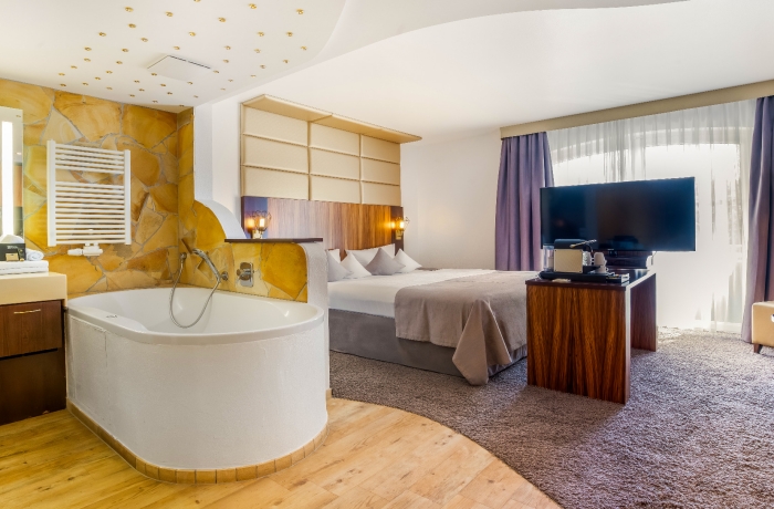 Das Hotel LIFESTYLE Resort Zum Kurfürsten präsentiert ein stilvolles Zimmer mit einer durchdachten Raumaufteilung: Das Bild zeigt verschiedene Bereiche des Zimmers, beginnend mit einem eleganten Waschbecken, das von einem großzügigen Unterschrank und einem beleuchteten Beauty-Spiegel flankiert wird. Neben diesem Bereich befindet sich eine einladende Whirlwanne, in der sprudelndes Wasser Entspannung verspricht. Durch eine geschickt platzierte halbhohe Mauer abgetrennt, lädt ein gemütliches Bett mit warmfarbigen Textilien zu erholsamen Nächten ein.