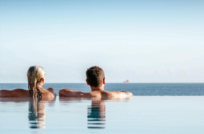 Zwei Personen entspannen in einem Infinitypool mit weitem Blick auf das ruhige Meer und den klaren Horizont.