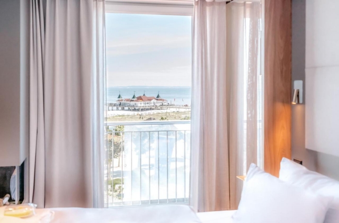 Helles Hotelzimmer mit bodentiefem Fenster und Balkon, das einen freien Blick auf Strand, Seebrücke und die Ostsee bietet.