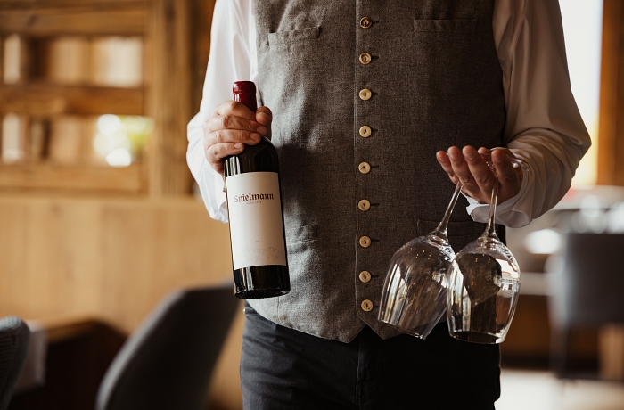 Ein Mann trägt eine Flasche Wein mit Hotel-Spielmann-Label sowie zwei Weingläser zu Hotelgästen