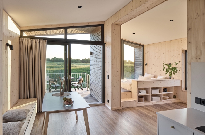 Ostsee Baumhaushotel: Helles Baumhaus-Loft mit nachhaltiger Holzarchitektur, bodentiefen Panoramafenstern und offenem Wohnkonzept. Die stilvolle Einrichtung, natürliche Materialien und die private Terrasse mit Blick auf die umliegende Wiesenlandschaft schaffen eine ruhige und naturnahe Atmosphäre für einen erholsamen Aufenthalt an der Ostsee.