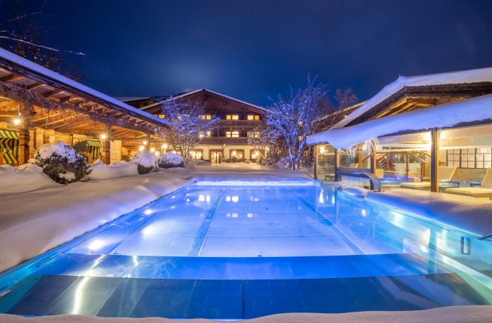 Im Fokus des Bildes ist ein beleuchteter Pool der umgeben von Schnee ist. der Pool si in einem Innenhof und  die Gebäude des Hotels sind rund herum.