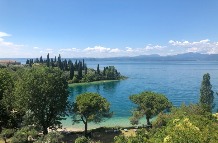 Blauer See und Hügel im Hintergrund. Im Vordergrund ein kleiner Sandstrand und grüne Bäume.