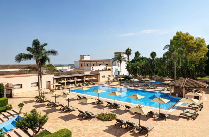 Mediterraner Hotelpool mit Palmen, Sonnenschirmen und Liegen.