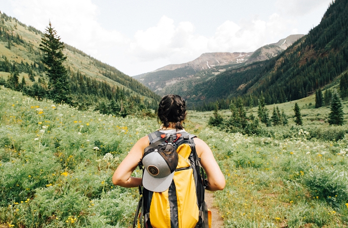 Eine Wanderin mit gelben Rucksack, an dem eine Kappe befestigt ist, schaut auf eine blumen- und grasbewachsene Wiese, im Hintergrund erstrecken sich Tannenwälder und Berge.