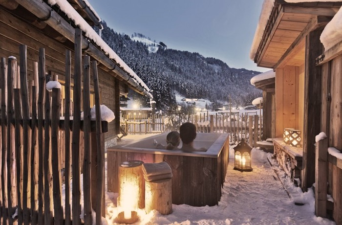 Schneebedeckter Bereich zwischen zwei Chalets mit einer Holzbadewanne umgeben von Laternen mit Waldlandschaft Hintergrund