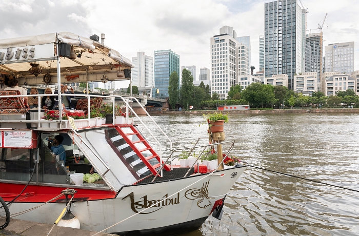 Zu sehen ist das Dönerboot auf dem Main, im Hintergrund das Mainufer, von Hoch- und Bürohäusern gesäumt