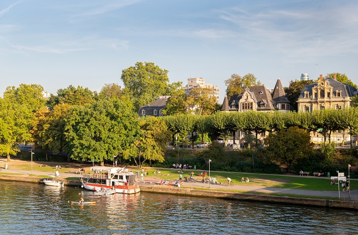 Geheimtipp Frankfurt: Blick auf das Mainufer mit Boot angelegt am Kai, in Hintergrund und hinter einer dichten Reihe Alleenbegrünung, historische Villen