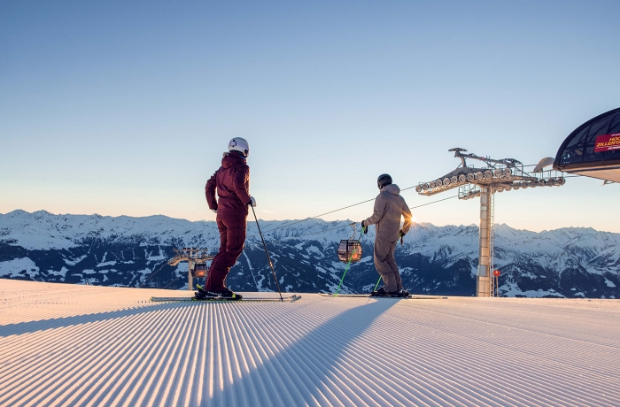 Zwei Skifahrer stehen an der frisch planierten Abfahrt und blicken auf das Bergpanorama. Hinten rechts ist der Pfeiler einer Seilbahn zu erkennen