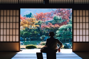 Ein Mann blickt im Schneidersitz mit Rücken zum Fotografen aus einem traditionellen, japanischen Haus mit Papier-Schiebetüren in einen üppig angelegten Garten
