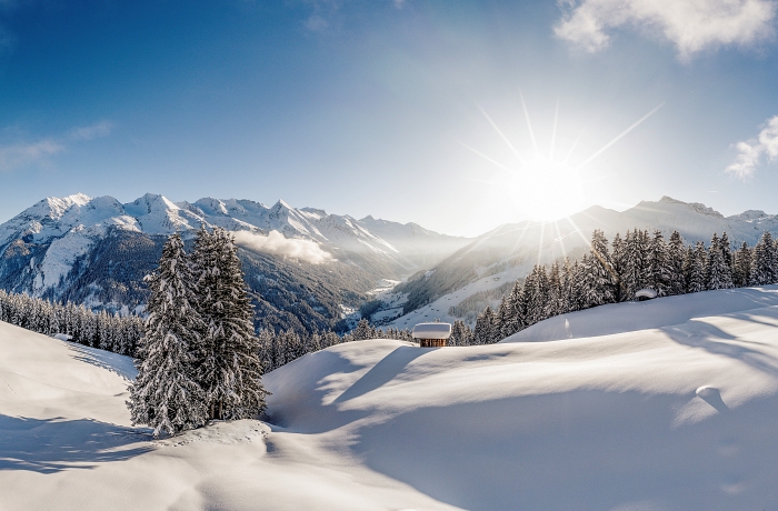 Verschneites Bergpanorama mit einer kleinen Holzhütte in der Mitte, umrahmt von verschneiten Tannen.