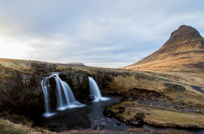 Ein kleiner Wasserfall fließt über Felsen in eine dunkle Lagune, im Hintergrund ragt ein markanter, pyramidenförmiger Berg in einer kargen Landschaft auf.