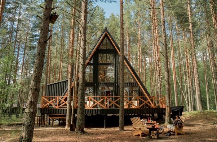 Ein modernes A-Frame-Holzhaus steht erhöht zwischen hohen Kiefern, davor sitzen zwei Personen an einer Feuerstelle auf Holzstühlen
